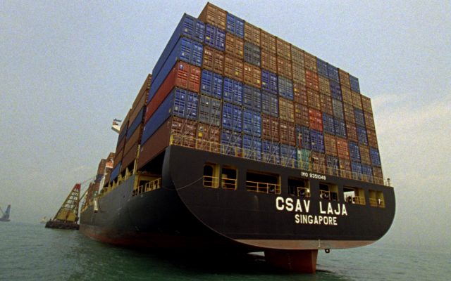 A huge ship laden with many shipping containers of different colours, viewed at an angle from behind.