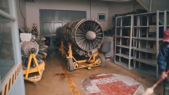 A dusty, stripped down airplane engine sits atop a wheeled yellow metal frame in a storage room. Empty metal shelves line the wall to the right, and another airplane component sits on another yellow frame to the left. On the floor in front of the engine, a large peeling red and white sticker reads 'SALIDA EXIT' with an arrow pointing forwards. Half out of frame, a man in blue overalls and a red cap sweeps the floor.