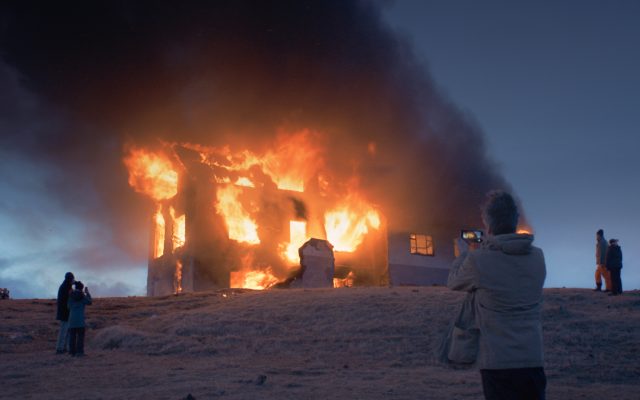 Against a clear, dusky sky, a large solitary farmhouse is engulfed in flames. A dark black smoke plumes upwards and leans left, blown by the wind. Dotted across the frozen hillside in the fore and middle ground are several onlookers. The nearest, a solitary man in a pale jacket, stands centre right with his back to us, taking a photo of the burning house on his smartphone.