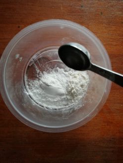 A round clear plastic yoghurt pot seen from above on a wooden surface. In the tub is some flour and some baking powder. A tablespoon measure hovers above the tub, pouring some water over the mixture.