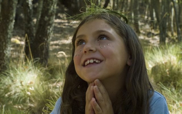 The close-up face of a young white girl with long brown hair with the backdrop of a sun dappled forest. The girl is grinning and clasping her hands beneath her chin in a prayer position, looking up and to the left as if in a daydream. Beneath her eyes she has stuck pretend grass seeds as if they are tears, and on her head she wears a halo made from grass.