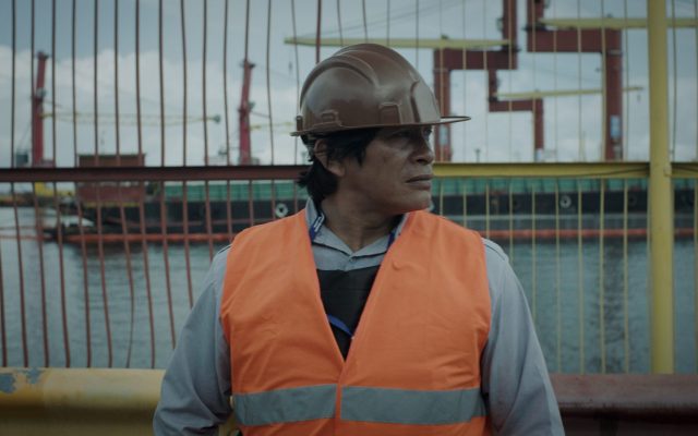 An indigenous Brazilian man stands centre frame, visible from the elbows up. He looks to the left and wears a high vis orange vest over a pale blue shirt and a brown hard hat. Behind him is a wire fence, with an industrial port beyond.