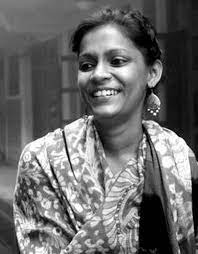 A black and white portrait of Ekta Mittal. Ekta is visible from the elbows up and is looking downwards to the left and smiling. She wears her dark hair pulled back with leaf shaped earrings and a patterned top