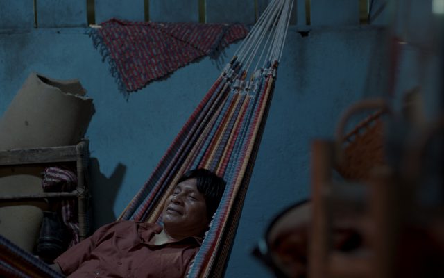 A man of indigenous Amazonian heritage sits in a stripy hammock within a domestic room painting a bright blue. The man has is eyes closed and is smiling.