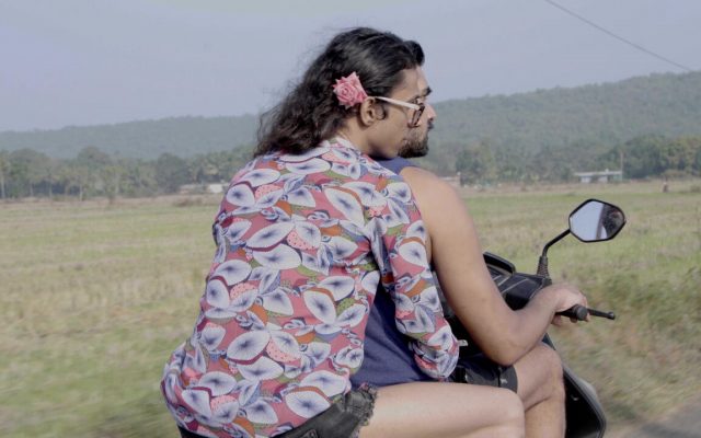 Two people riding a motorcycle through the countryside, viewed from slightly behind and to the right of them. The person driving the motorcycle has short dark hair and wears a sleeveless dark blue vest. The passenger behind has flowing long dark wavy hair with a pink rose pin behind the right ear. They wear shorts and a colourful pink and blue patterned shirt. They have their arms wrapped around the waist and their chin resting upon the shoulder of the person in front.