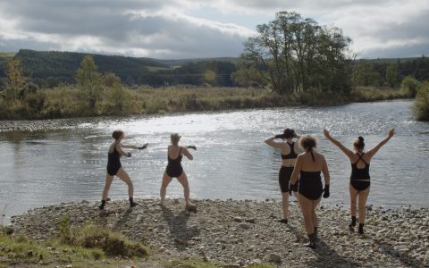 A group of five people in black swimming costumes are preparing to swim in the river. They stand on a small gravel beach in front of a serene river pool, bathed in sunlight. Beyond there is a green field, some trees and rolling mountains in the distance. The swimmers and doing stretches and warm ups.
