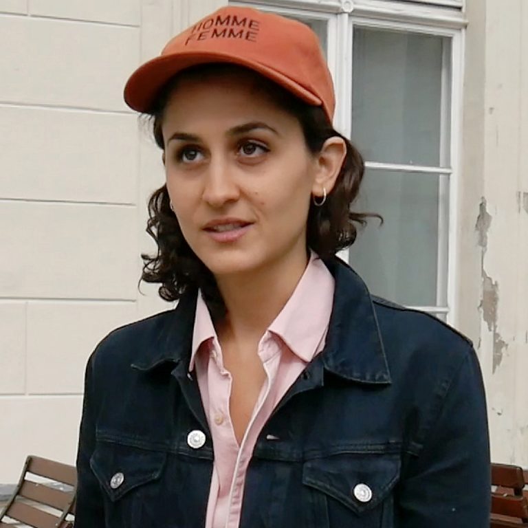 A woman with shoulder length dark curly hair, wearing a pink buttoned short, a dark denim overshirt, a ochre coloured cap and small silver loop earrings. The cap has black writing that reads 'HOMME FEMME'. Behind her is an off-white brick building with some wooden outdoor seating.