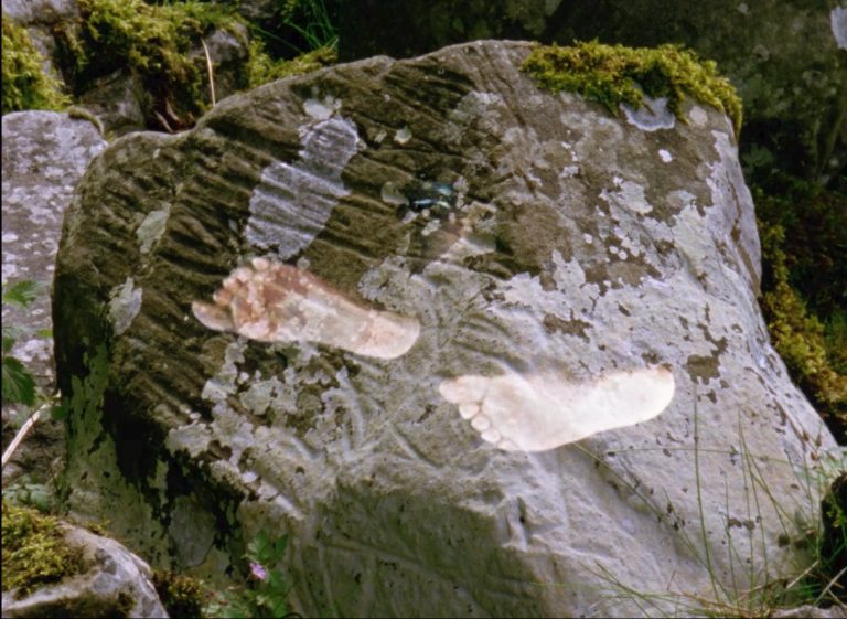 A large boulder encrusted with lichen and moss, superimposed with an image of two bare feet in a walking motion, as if they are leaving footprints over the boulder.