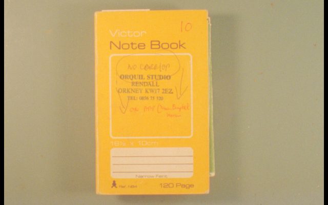 A yellow notebook sitting on a table, viewed from directly above. The notebook is a vintage style and has various text printed on it including an Orkney address and telephone number, along with scribbled handwriting in red pen.