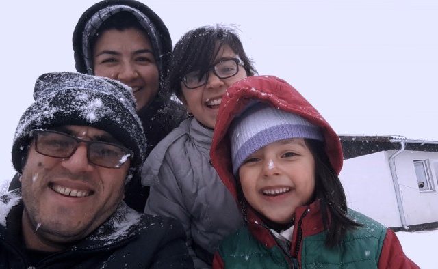 A family take a selfie in the snow. They smile gleefully, as if snow is novel to them. In the background is a single storey white building.