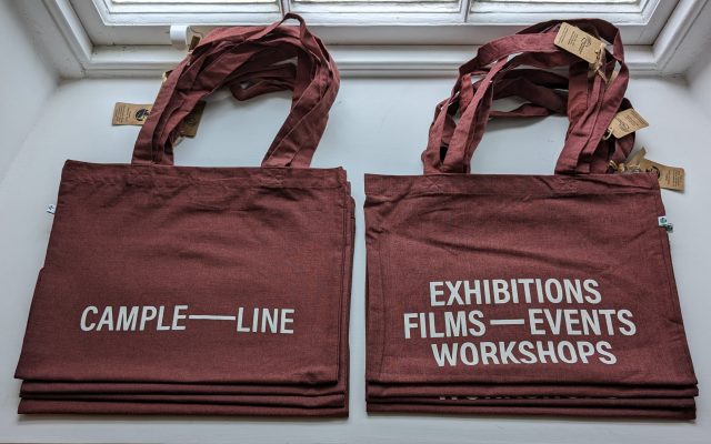 Cloth tote bags in a dark red colour are folded neatly on a bright window ledge. The bags have block lettering that say "CAMPLE LINE' on one side and 'EXHIBITIONS, FILMS, EVENTS, WORKSHOPS' in the other side.