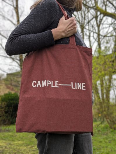 A woman holds a dark red cloth bag over her shoulder, with block lettering on the bag that reads 'CAMPLE-LINE'. Behind her there is trees and greenery, with the archway of a railway viaduct in the background.