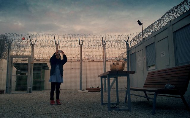 A young girl stands in the courtyard of a secure compound at sunset. There are high barbed wire fences behind her. She is taking a video on a mobile phone, pointing the camera up towards the sky.