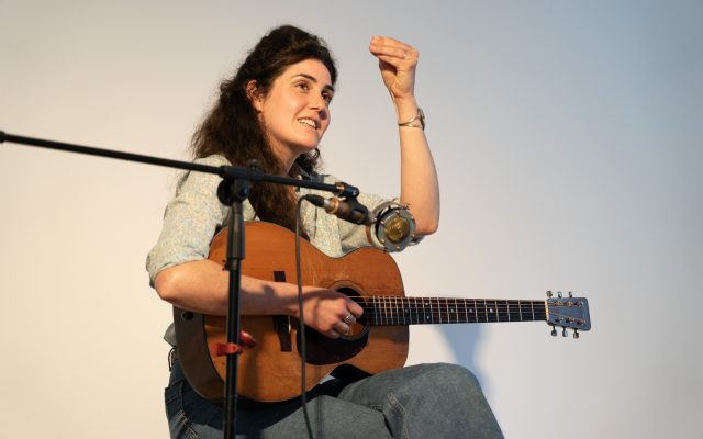 Musician Sarah Jane Scouten sits on a stool holding a guitar whilst chatting to the audience.