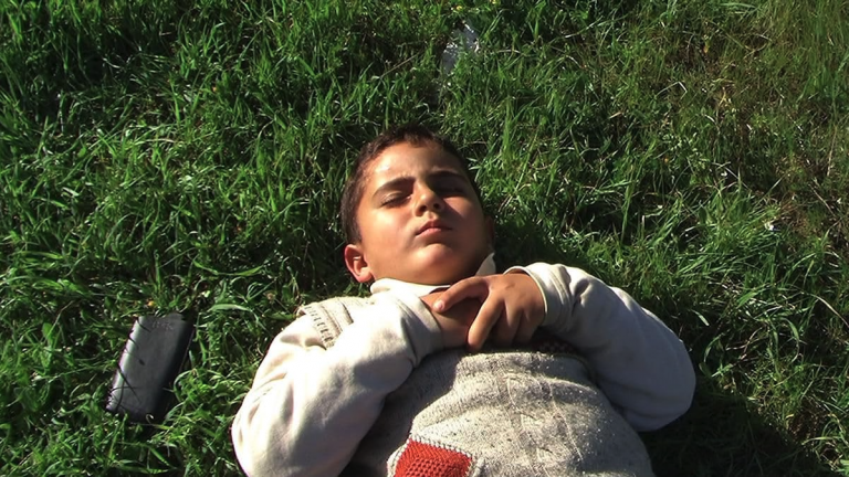 A young boy lies on grass in the sunshine with his eyes closed. His hands are clasped together on his chest and there is a mobile phone on the grass beside him.