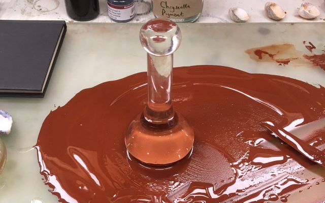A workspace with watery iron oxide pigment smeared on a mat. There is a glass muller - an upright handheld glass tool used for grinding pigment - standing in the centre.
