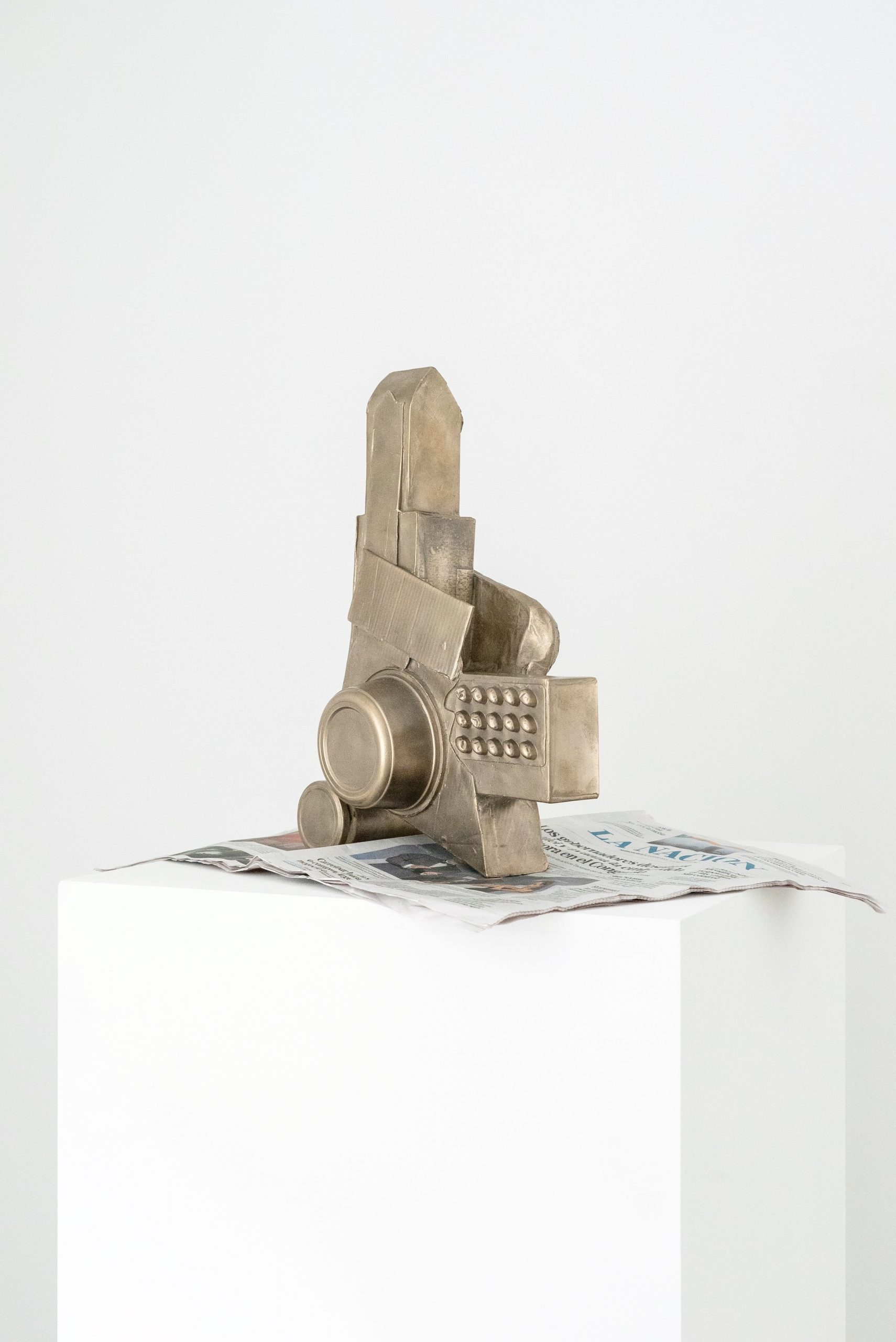 Amalia Pica’s brone sculpture, ‘Another One’s Treasure’ (2025) rests on a newspaper on top of a white stand. It is roughly a hand and a half high, and half a hand wide, and is made from junk metal found around the home. It appears to have a tall, pointed head, short arms, and its lower body is made from tins and a cheese grater.