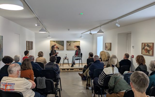 Bryony Rose and Tina Fiske sit on a raised platform in the downstairs gallery in conversation. In front of them, rows of seated people listen and watch.