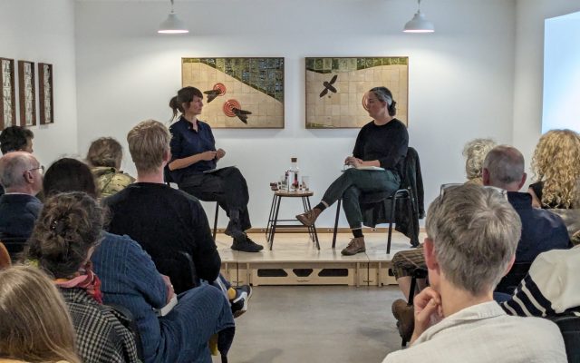 Lotte Gertz and Amy Winstanley sit in conversation on a raised platform in the downstairs gallery. Rows of people are seated to listen to the talk.