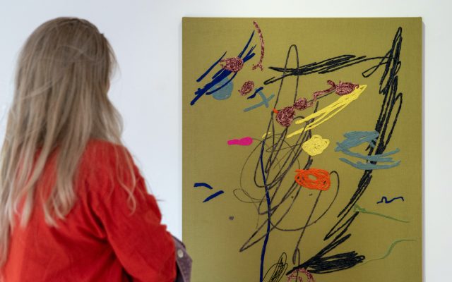 An art piece from the exhibition, ‘Keepsake’ by Amalia Pica, is hung in the upper gallery, being viewed by a gallery visitor. It is roughly the size of a large beach towel, and is hung vertically. The backdrop is golden, and freeform patterns reminiscent of a child’s drawing are stitched on top in both black and coloured cotton and wool.
