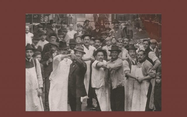 The book cover for ‘Milongas’ by Edgardo Cozarinsky, translated from Spanish by Valerie Miles. The cover is burgundy and displays a black and white photograph in the middle. In the image, a crowd of men stand in the street looking at the camera. Those at the front stand in a dancing hold, with clasped hands stretched out towards the camera. White text displays the title, author’s name and translator.