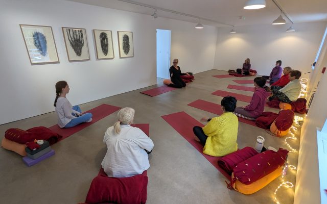 Red yoga mats are laid out in the downstairs gallery in a semi-circle. Jane Bailey gives a talk to gallery visitors who sit cross-legged on the mats.