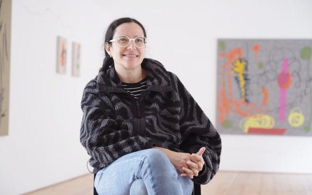 A photograph of Amalia Pica in the upper gallery smiling at someone out of frame. Artwork from her collection, ‘Keepsake’, is displayed on the walls behind her.