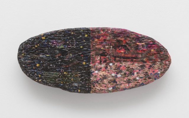 The sculpture ‘Medallion’ (2025) by Anderson Borba. A pill-shaped sculpture where the left half is black and the right half is pink, with many different colours layered in, creating an iridescent and oily appearance. The surface is very textured and appears scaly and rippling. Faint distorted images can be made out on the sculpture’s surface.