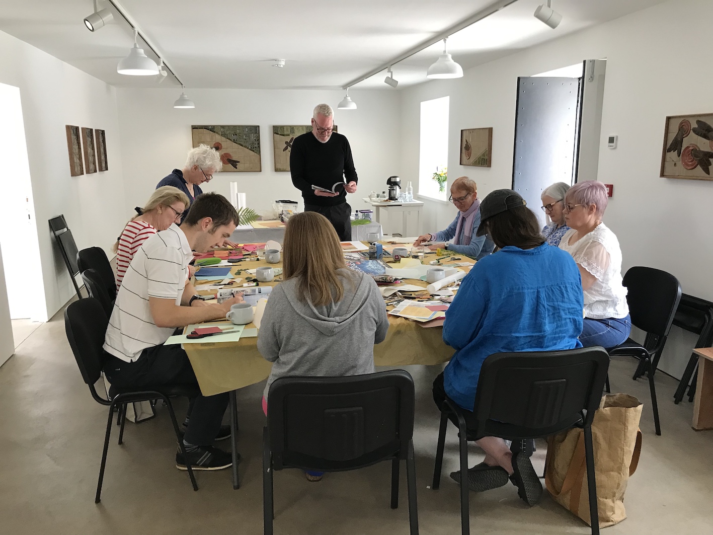 Frank Hayes leads a drawing workshop at CAMPLE LINE. 8 people sit around a large table covered with art materials, each absorbed in their own creative process. Frank stands at the end of the table, working on his own drawing. Bright sunlight streams in to the room through the doors and windows.