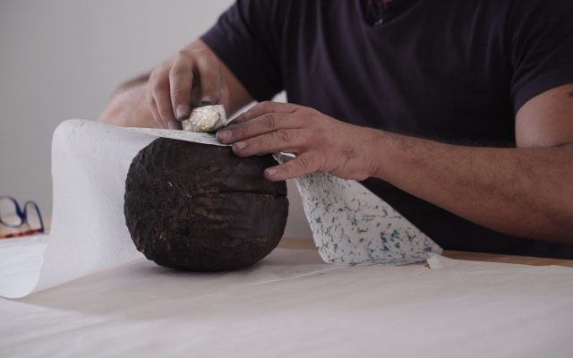 A close up shot of hands creating a wood rubbing or 'frottage'. The image shows a large piece of carved dark wood roughly the shape of a watermelon, with a piece of white paper placed on top. The person is rubbing another long, thin piece of carved wood, shaped a bit like a weaving shuttle and coated with coloured oil pastel, over the paper to pick up the textured patterns of the wood below.