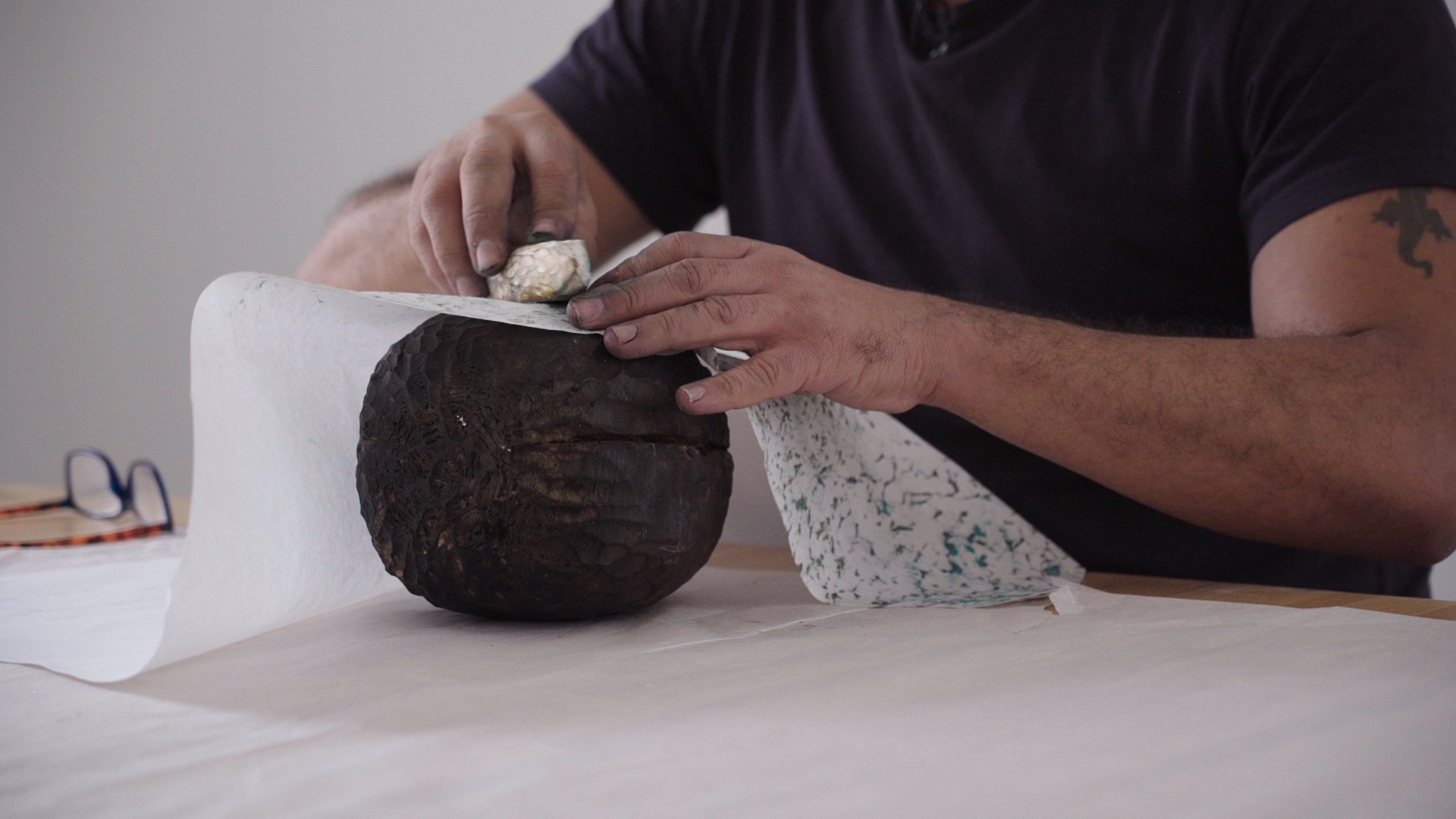 A close up shot of hands creating a wood rubbing or 'frottage'. The image shows a large piece of carved dark wood roughly the shape of a watermelon, with a piece of white paper placed on top. The person is rubbing another long, thin piece of carved wood, shaped a bit like a weaving shuttle and coated with coloured oil pastel, over the paper to pick up the textured patterns of the wood below.