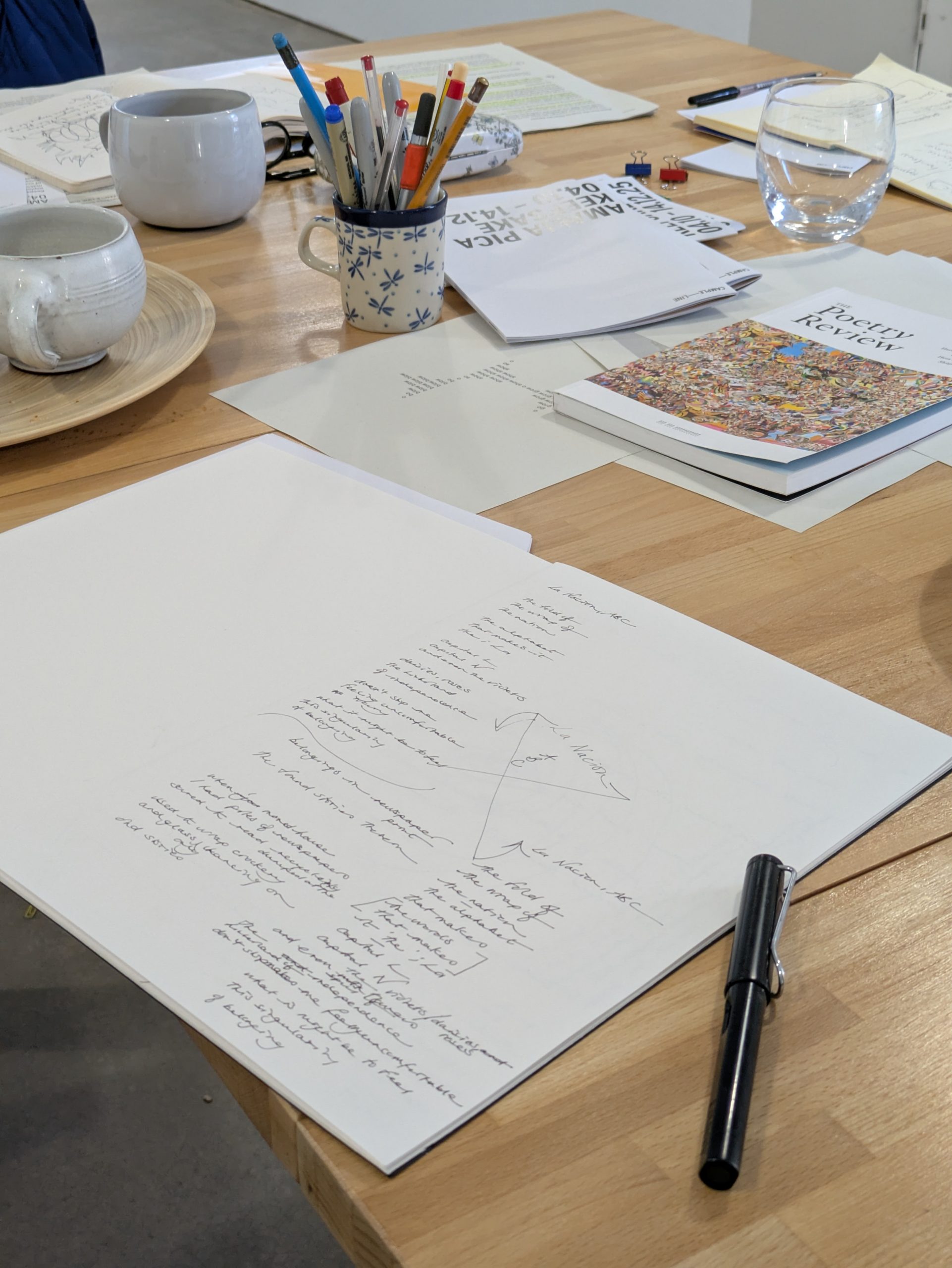 A notebook sits open scrawled with lines of text and fragments of poetry. It sits on a table surrounded by other publications, writing materials and cups of tea.