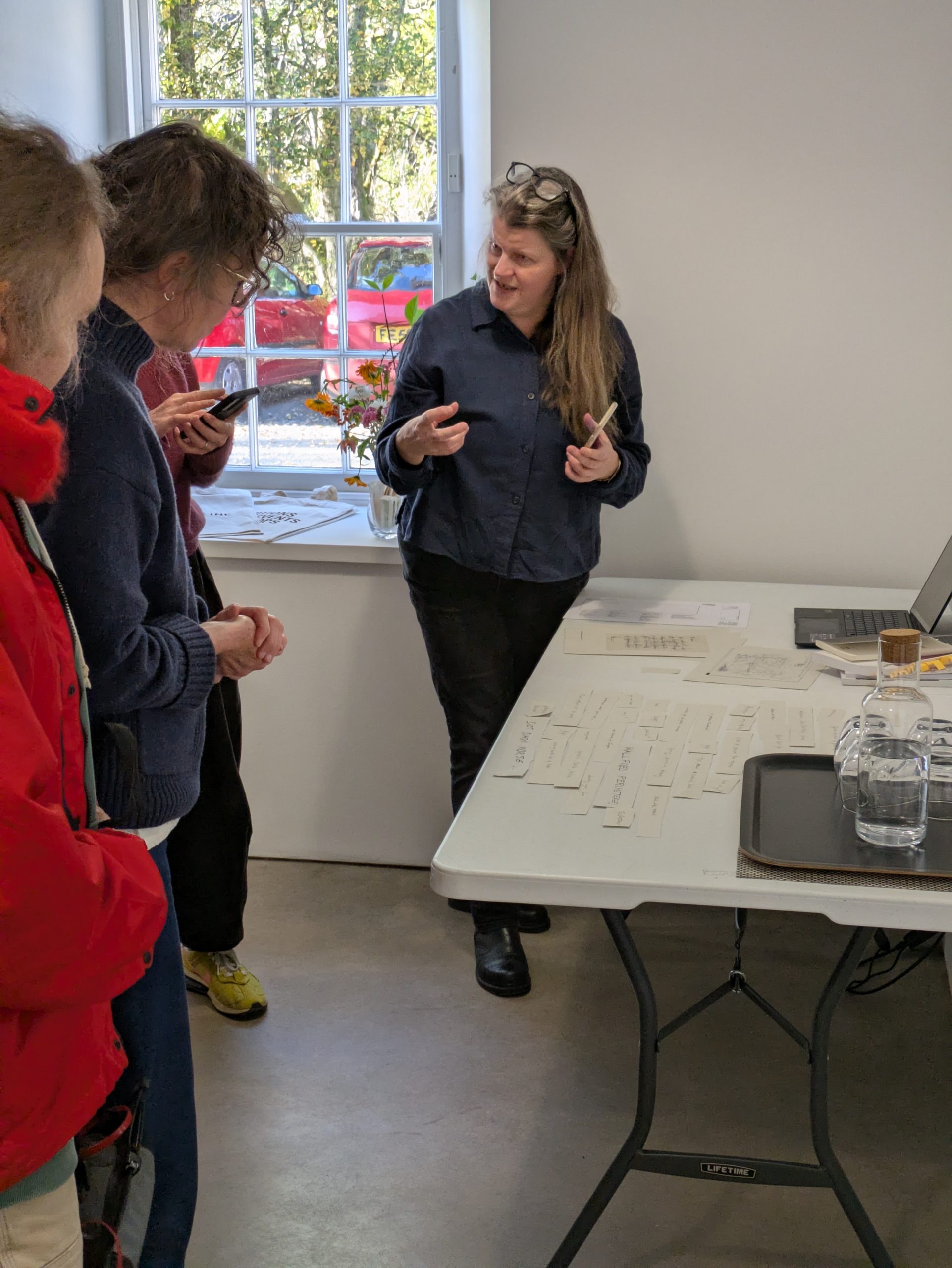 Writer Carolyn Hashimoto leads a creative writing workshop at CAMPLE LINE. Participants gather around a table to look at fragments of collage poetry, as Carolyn explains the technique.