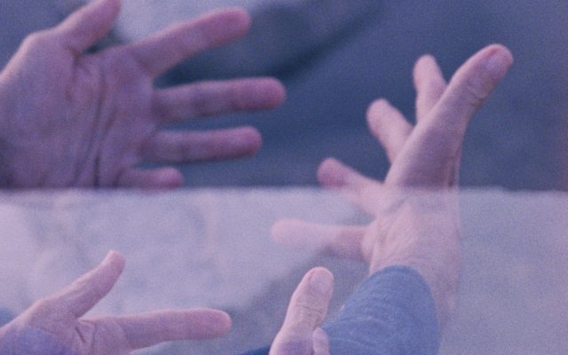 A grainy, double-exposed 16mm image of two pairs of hands making the same gesture, outstretched with palms open and facing slightly upwards. Blue and pink colour tones suggest the image is shot in the evening, close to dusk.