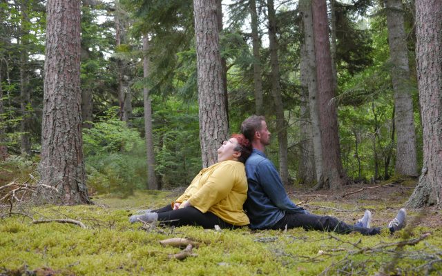 Two people seen in profile sit back to back on a mossy forest floor, surrounded by the trunks of tall fir trees. A man in a denim shirt and jeans sits on the right and gazes into the woods. A woman with short red hair and wearing a yellow top sits on the left and gazes up towards the sky, resting the top of her head on the back of the man's neck.