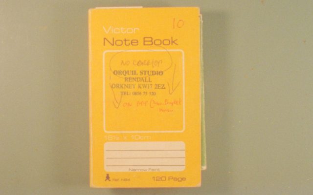 A yellow notebook sitting on a table, viewed from directly above. The notebook is a vintage style and has various text printed on it including an Orkney address and telephone number, along with scribbled handwriting in red pen.