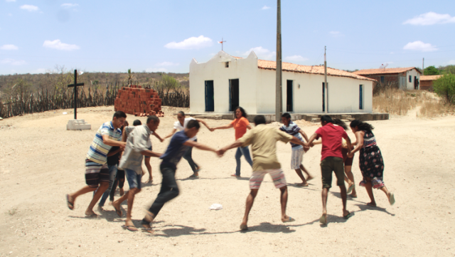 On a bright sunny day, a group of men and women hold hands and run in a circle anti-clockwise. They wear shorts and t-shirts in various colours. The ground beneath their bare feet is bare and sandy. In the background there is a long, single storey white brick building with a beige coloured roof. The sky is blue with some tiny, wispy white clouds.